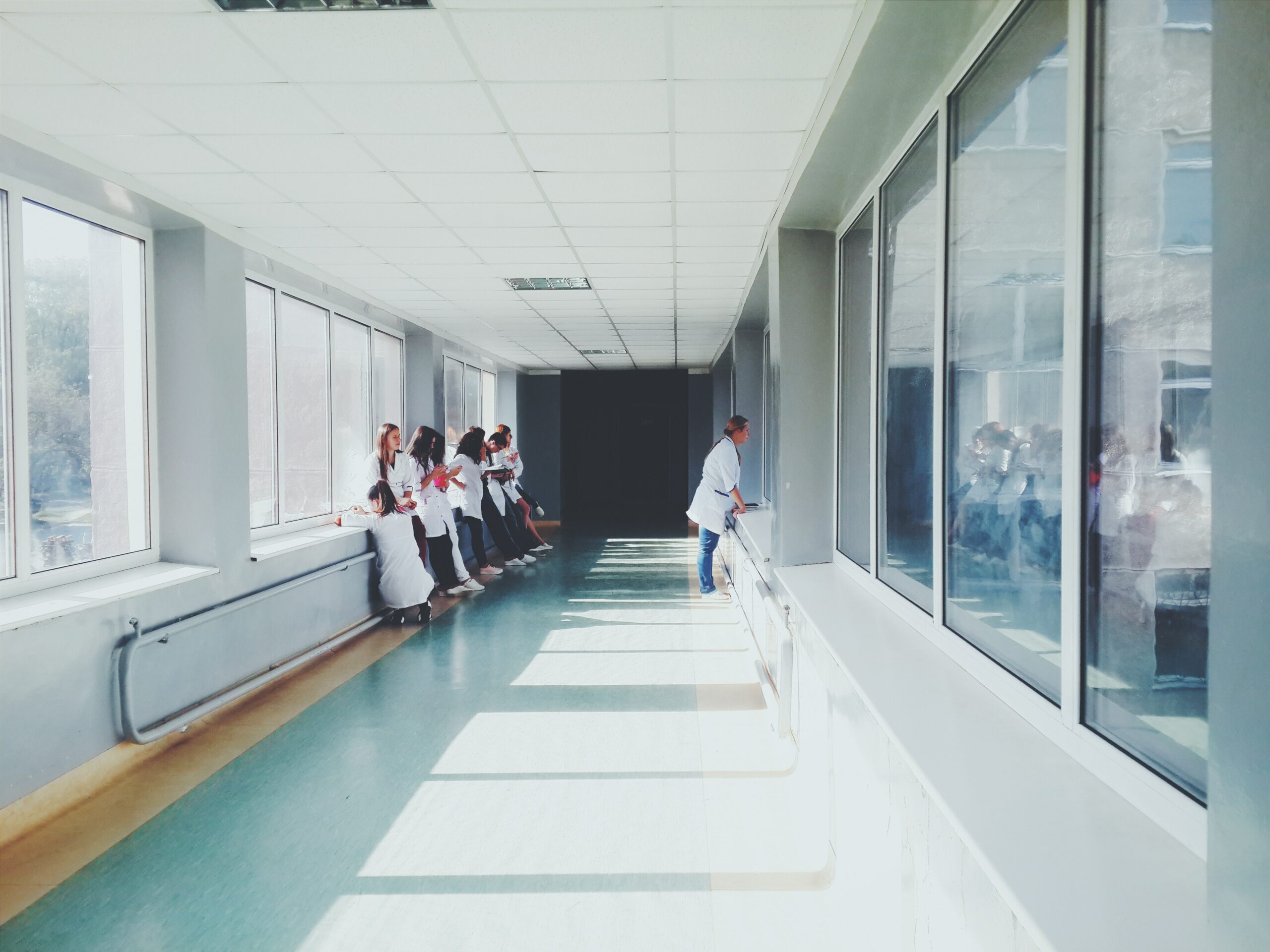 Long hallway in a hospital. One side has floor to ceiling windows and the other has doctors learning against the wall.